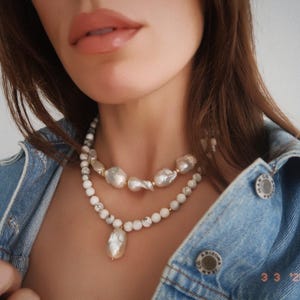 May include: A woman wearing a denim jacket with two layered necklaces. The top necklace is made of white pearls and the bottom necklace is made of white and gray stones. The bottom necklace has a large white pearl pendant.