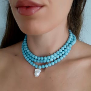 May include: A multi-strand necklace featuring light blue beads. A large, irregularly shaped white pearl pendant hangs from the necklace. The necklace is worn around the neck.