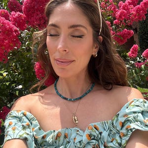 May include: A woman wearing a light blue off-the-shoulder top with an orange floral pattern. She is wearing a turquoise beaded necklace and a gold necklace with a fish pendant. Sunglasses are on top of her head, and pink flowers are in the background.