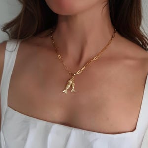 Könnte beinhalten: Eine goldene Kettenkette mit einem Anhänger mit zwei kleinen goldenen Fisch-Anhängern. Die Kette wird von einer Person mit langen braunen Haaren getragen und ist im Bild zentriert.