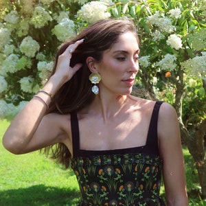 May include: A woman with long brown hair wearing a black and green floral dress with a black bodice. She is wearing large gold and pearl earrings.