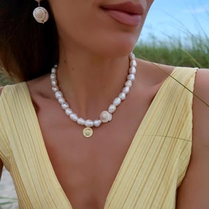Puede incluir: Se muestra un collar de perlas y pendientes a juego. El collar tiene perlas grandes e irregulares, un adorno de concha y un pequeño colgante dorado. Los pendientes tienen un diseño similar, con una combinación de perlas y conchas.