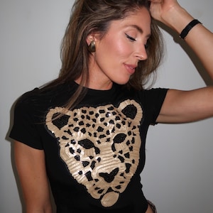 May include: Black t-shirt featuring a gold sequined leopard face design. The shirt has short sleeves and a crew neck. The model is wearing gold earrings and a black bracelet. The pants have a leopard print.