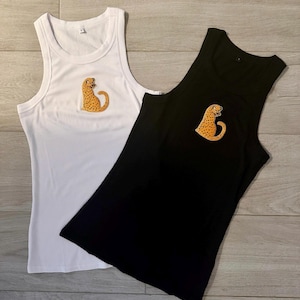 May include: Two ribbed tank tops, one white and one black, each featuring a small embroidered patch of a yellow leopard with black spots on the upper chest area. The sleeveless garments have a round neckline and a fitted, textured fabric design.