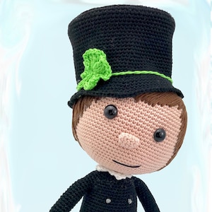 Może przedstawiać: Lalka szydełkowa w czarnym cylindrze z zielonym listkiem, czarnej kurtce i czarnych spodniach. Lalka ma brązowe włosy szydełkowe i uśmiechniętą twarz.