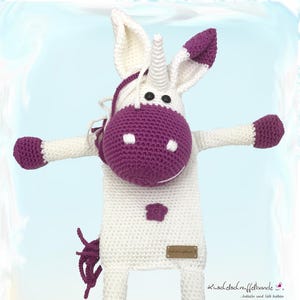 Può includere: Un unicorno bianco all'uncinetto con dettagli viola. L'unicorno ha un corno lungo, un naso viola e zoccoli viola. L'unicorno tiene un piccolo fiore viola nella sua mano. Il testo "kuschel-stoff-bänder" è su un'etichetta marrone attaccata all'unicorno.