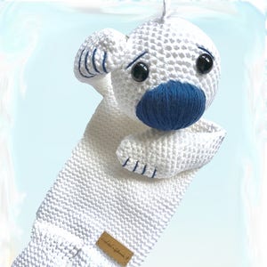 Può includere: Un animale di peluche bianco fatto a mano con naso e dettagli blu. Il giocattolo ha grandi occhi neri e una piccola etichetta di legno. Il testo "Kuschelschnuffelbande...häkeln und lieb haben" è visibile.