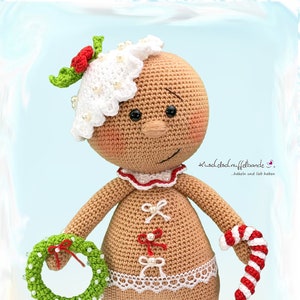 Puede incluir: Una muñeca de jengibre de crochet que lleva un gorro blanco de crochet con bayas rojas, una falda de encaje blanco de crochet y sostiene una corona verde y un bastón de caramelo rojo y blanco.