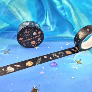 May include: Black washi tape featuring a repeating pattern of pumpkins, leaves, mushrooms, and a cat. The tape roll is labelled "ellyn art" and "Pumpkin Patch". The tape is unrolled, displaying the design. The background is a blue fabric with star-shaped confetti.