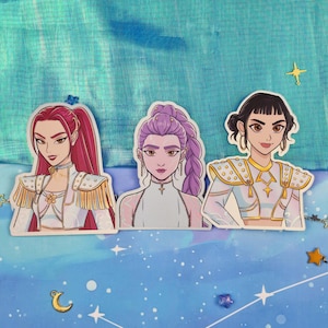 Könnte beinhalten: Drei Aufkleber mit stilisierten Illustrationen von Frauen mit unterschiedlichen Frisuren und Outfits. Die erste Frau hat rotes Haar, die zweite hat lila Haar und die dritte hat schwarzes Haar. Die Aufkleber befinden sich auf einem blauen Hintergrund mit Stern- und Monddekorationen.