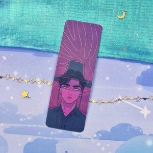 May include: A bookmark featuring an illustration of a person wearing a hat. The artwork has a purple and pink color scheme, with the person's eyes glowing yellow. The background is a gradient of purple and pink lines.