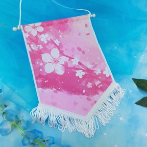 May include: A pink and white fabric banner with a watercolour design of cherry blossoms and white fringe. The banner is hanging from a white string with wooden dowels at the top.