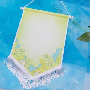 Könnte beinhalten: Ein weißes Banner mit einem gelben und grünen Blumendesign. Das Banner hat einen weißen Fransenrand und hängt an einer weißen Schnur.