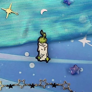 Pode incluir: Um broche de esmalte de uma vela branca com uma chama verde sobre um fundo azul com estrelas douradas e uma corrente dourada.