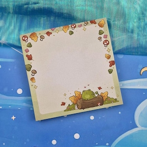 Op de afbeelding: Een groene kikker die op een boomstam zit met een paddenstoel en bladeren eromheen. De achtergrond is lichtblauw met witte sterren. De kikker bevindt zich op een notitieblok met een witte achtergrond en een groene rand. De rand is versierd met paddenstoelen, bladeren en harten.