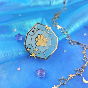 May include: A gold-tone enamel pin shaped like a blue gemstone with a gold paw print inside. The pin is surrounded by gold stars and is on a blue background.