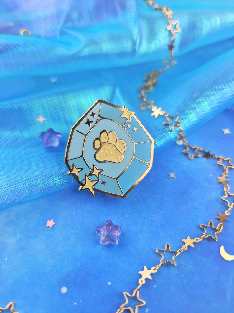 May include: A gold-tone enamel pin shaped like a blue gemstone with a gold paw print inside. The pin is surrounded by gold stars and is on a blue background.