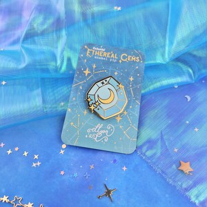 May include: A blue and gold enamel pin featuring a celestial gem with a crescent moon inside. The pin is on a card with the text "Celestial Ethereal Gems enamel pin" and "ellyon art".