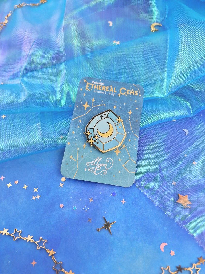 May include: A blue and gold enamel pin featuring a celestial gem with a crescent moon inside. The pin is on a card with the text "Celestial Ethereal Gems enamel pin" and "ellyon art".