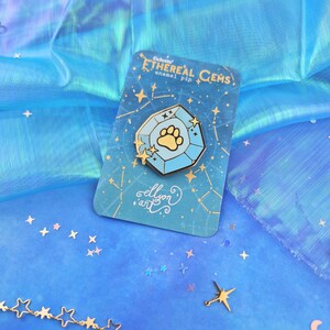 May include: A blue and gold enamel pin featuring a paw print inside a celestial gem. The pin is on a card with the text "Celestial Ethereal Gems enamel pin" and "ellison art".