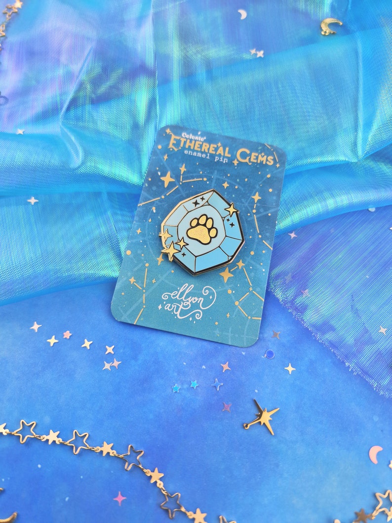 May include: A blue and gold enamel pin featuring a paw print inside a celestial gem. The pin is on a card with the text "Celestial Ethereal Gems enamel pin" and "ellison art".