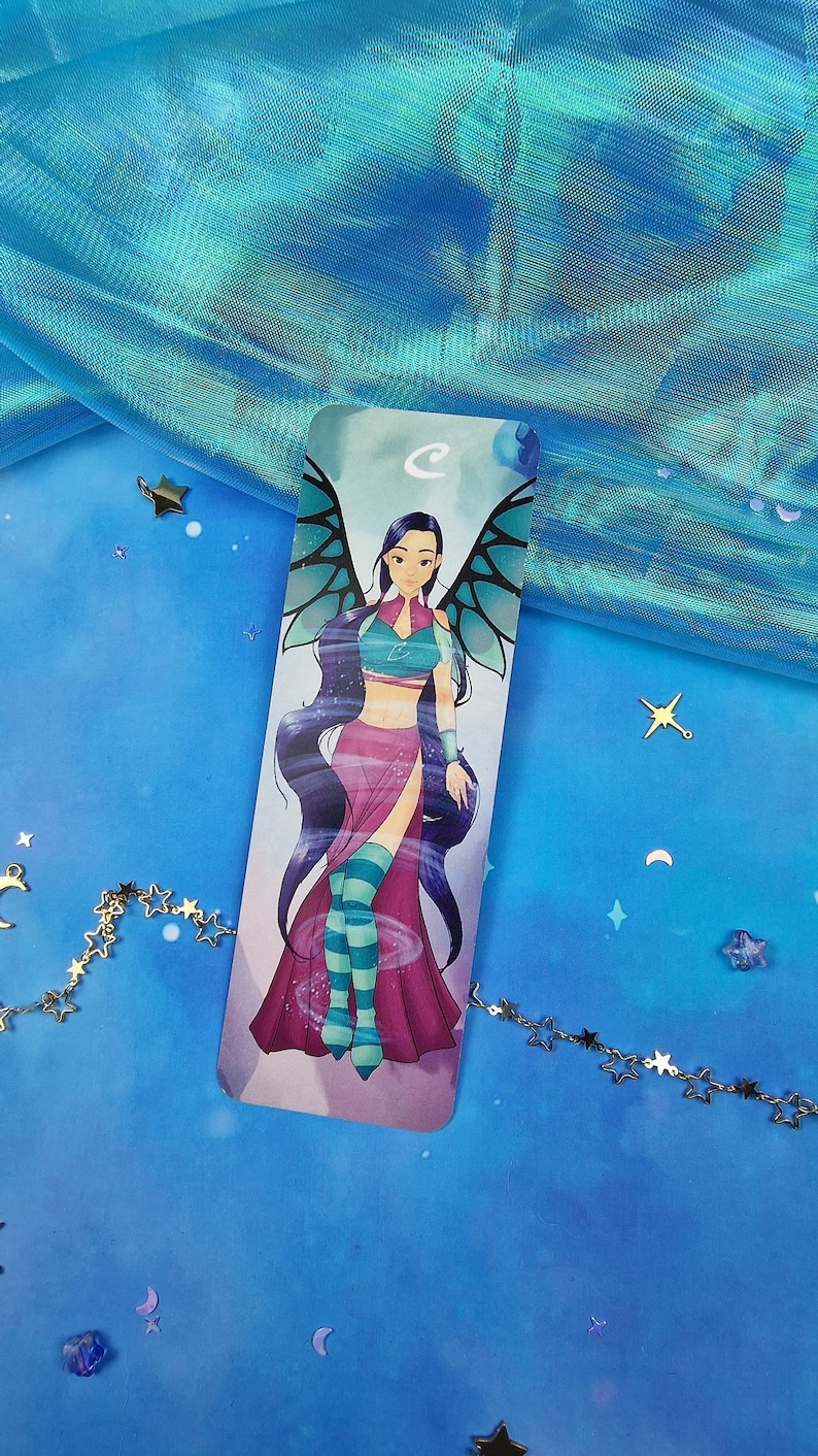May include: A bookmark featuring a cartoon illustration of a fairy with long purple hair, wearing a blue and pink outfit with wings. The fairy is standing on a swirling blue background.