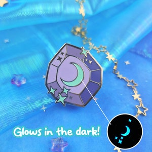 May include: A purple and blue enamel pin shaped like a gemstone with a crescent moon and stars inside. The pin glows in the dark, as shown in the image.