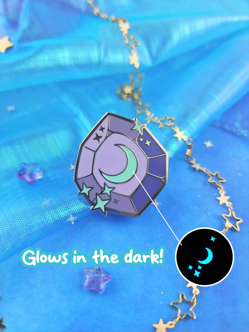 May include: A purple and blue enamel pin shaped like a gemstone with a crescent moon and stars inside. The pin glows in the dark, as shown in the image.