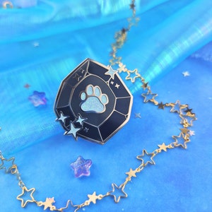 May include: A black and blue enamel pin shaped like a gemstone with a white paw print in the center and white stars around the edge. The pin is on a blue background with a gold chain with star links.
