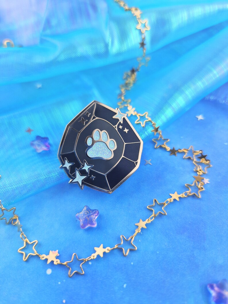 May include: A black and blue enamel pin shaped like a gemstone with a white paw print in the center and white stars around the edge. The pin is on a blue background with a gold chain with star links.