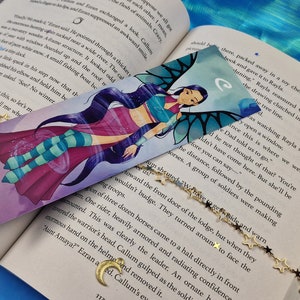 May include: A colourful bookmark featuring a cartoon character with blue hair, a pink and blue outfit, and wings. The character is surrounded by swirling purple and blue designs. The bookmark is on top of an open book with a gold chain with star charms.