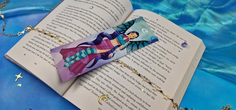 May include: A colourful bookmark featuring a cartoon character with blue hair, a pink and blue outfit, and wings. The character is surrounded by swirling purple and blue designs. The bookmark is on top of an open book with a gold chain with star charms.