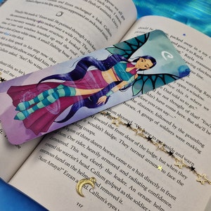 May include: A colourful bookmark featuring a cartoon character with blue hair and wings. The character is wearing a pink and blue outfit and is surrounded by a swirling purple and blue background. The bookmark has the letter "C" in the top right corner.