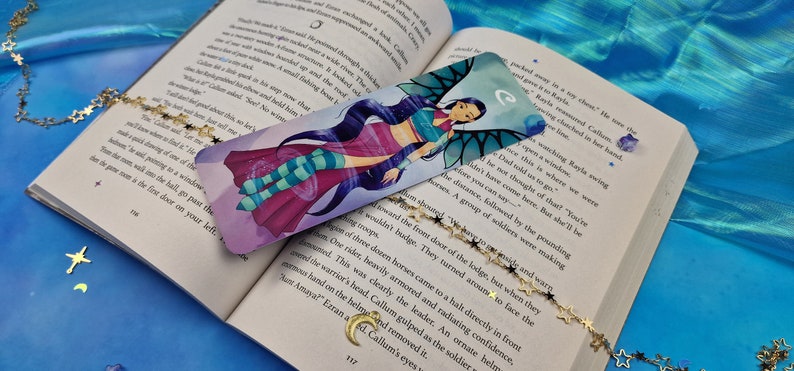May include: A colourful bookmark featuring a cartoon character with blue hair and wings. The character is wearing a pink and blue outfit and is surrounded by a swirling purple and blue background. The bookmark has the letter "C" in the top right corner.