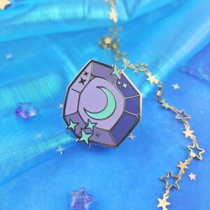 May include: A purple and blue enamel pin shaped like a gemstone with a crescent moon and stars inside. The pin is on a blue background with a gold chain of stars.