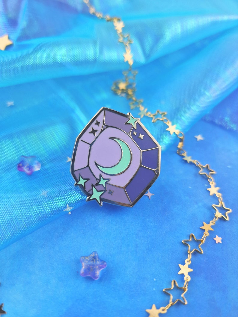 May include: A purple and blue enamel pin shaped like a gemstone with a crescent moon and stars inside. The pin is on a blue background with a gold chain of stars.