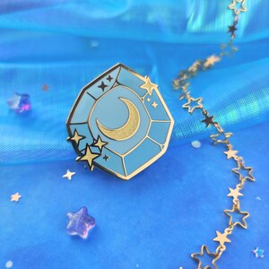 May include: A gold-tone enamel pin featuring a blue gemstone with a crescent moon and stars inside. The pin is on a blue iridescent background with a gold chain of stars.