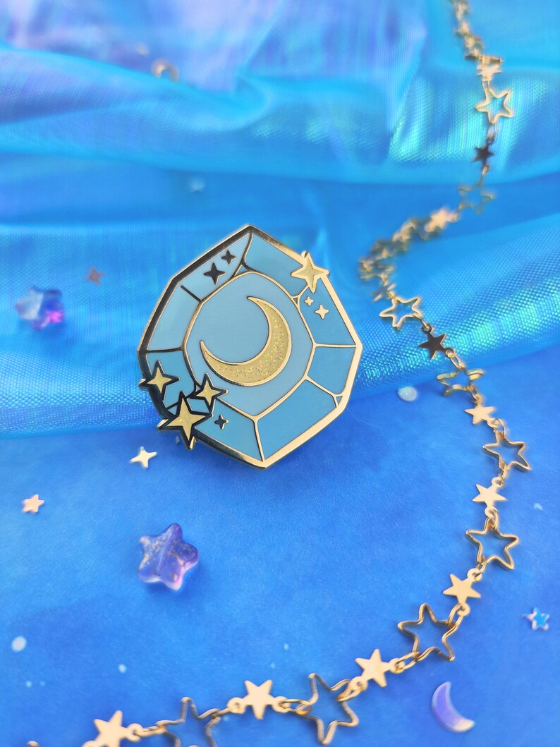 May include: A gold-tone enamel pin featuring a blue gemstone with a crescent moon and stars inside. The pin is on a blue iridescent background with a gold chain of stars.