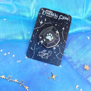 May include: A dark blue enamel pin shaped like a gemstone with a white paw print inside. The pin is on a card with a black background and white stars. The card says "Midnight Ethereal Gems enamel pin" and "ellison art".