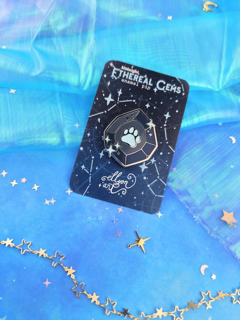 May include: A dark blue enamel pin shaped like a gemstone with a white paw print inside. The pin is on a card with a black background and white stars. The card says "Midnight Ethereal Gems enamel pin" and "ellison art".