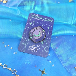 May include: A purple and blue enamel pin shaped like a gemstone with a crescent moon and stars inside. The pin is on a card with the text "Ethereal Gems enamel pin" and "ellyon art".