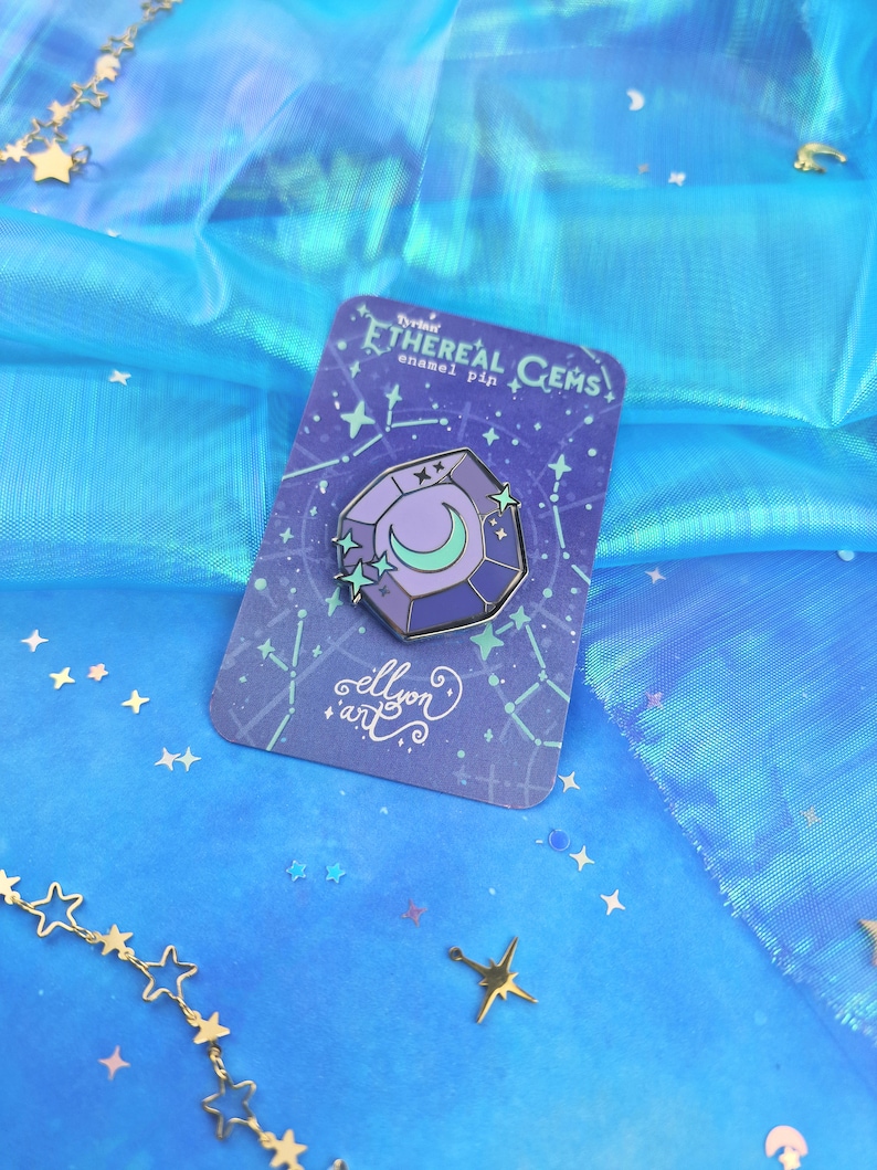 May include: A purple and blue enamel pin shaped like a gemstone with a crescent moon and stars inside. The pin is on a card with the text "Ethereal Gems enamel pin" and "ellyon art".