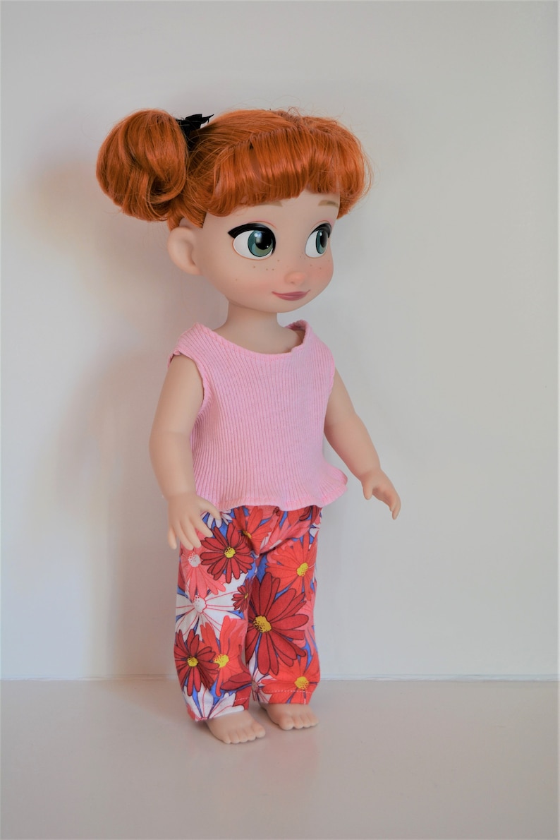 Puede incluir: Una mu&ntilde;eca de pelo rojo que lleva una camiseta rosa sin mangas y pantalones de estampado floral. La mu&ntilde;eca tiene ojos verdes y un lazo negro en el pelo.