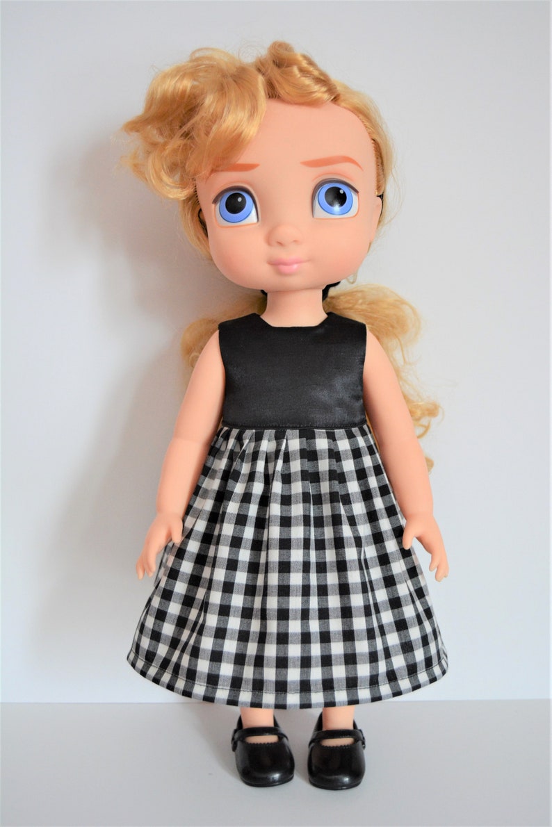 May include: A doll with blonde curly hair wearing a black and white gingham dress with a black satin bodice. The doll is wearing black shoes.