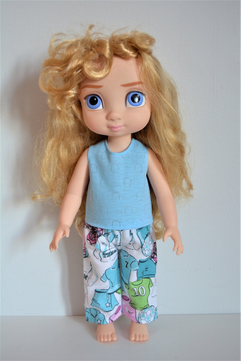 Puede incluir: Una mu&ntilde;eca con el pelo largo y rizado rubio que lleva una camiseta azul claro sin mangas con una falda estampada en blanco y azul. La mu&ntilde;eca tiene ojos azules y una boca rosa p&aacute;lida.
