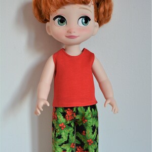 Puede incluir: Una mu&ntilde;eca de pelo rojo con una camiseta roja sin mangas y pantalones de estampado de acebo verde y rojo.
