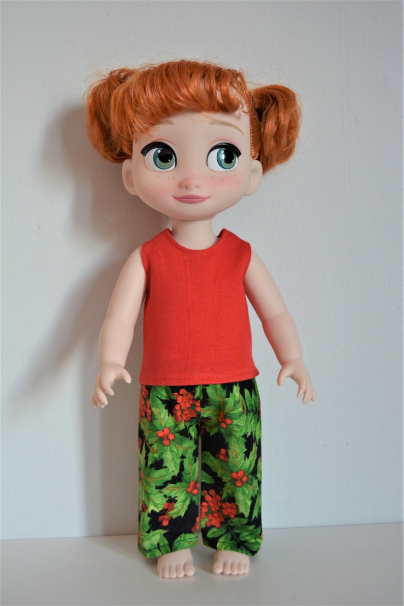 Puede incluir: Una mu&ntilde;eca de pelo rojo con una camiseta roja sin mangas y pantalones de estampado de acebo verde y rojo.