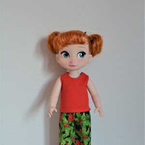 Puede incluir: Una camiseta roja sin mangas y pantalones de estampado de acebo verde y rojo para una mu&ntilde;eca. La mu&ntilde;eca tiene el pelo rojo y los ojos azules.