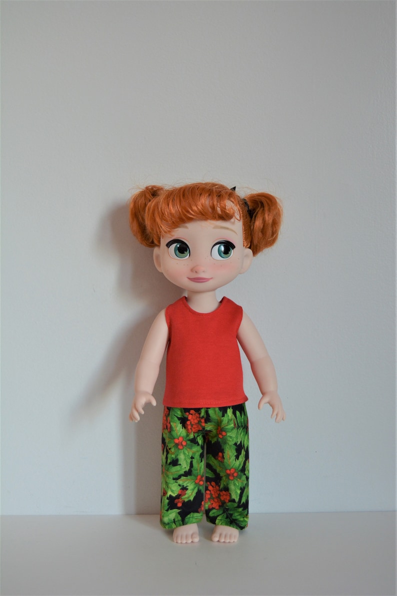 Puede incluir: Una camiseta roja sin mangas y pantalones de estampado de acebo verde y rojo para una mu&ntilde;eca. La mu&ntilde;eca tiene el pelo rojo y los ojos azules.