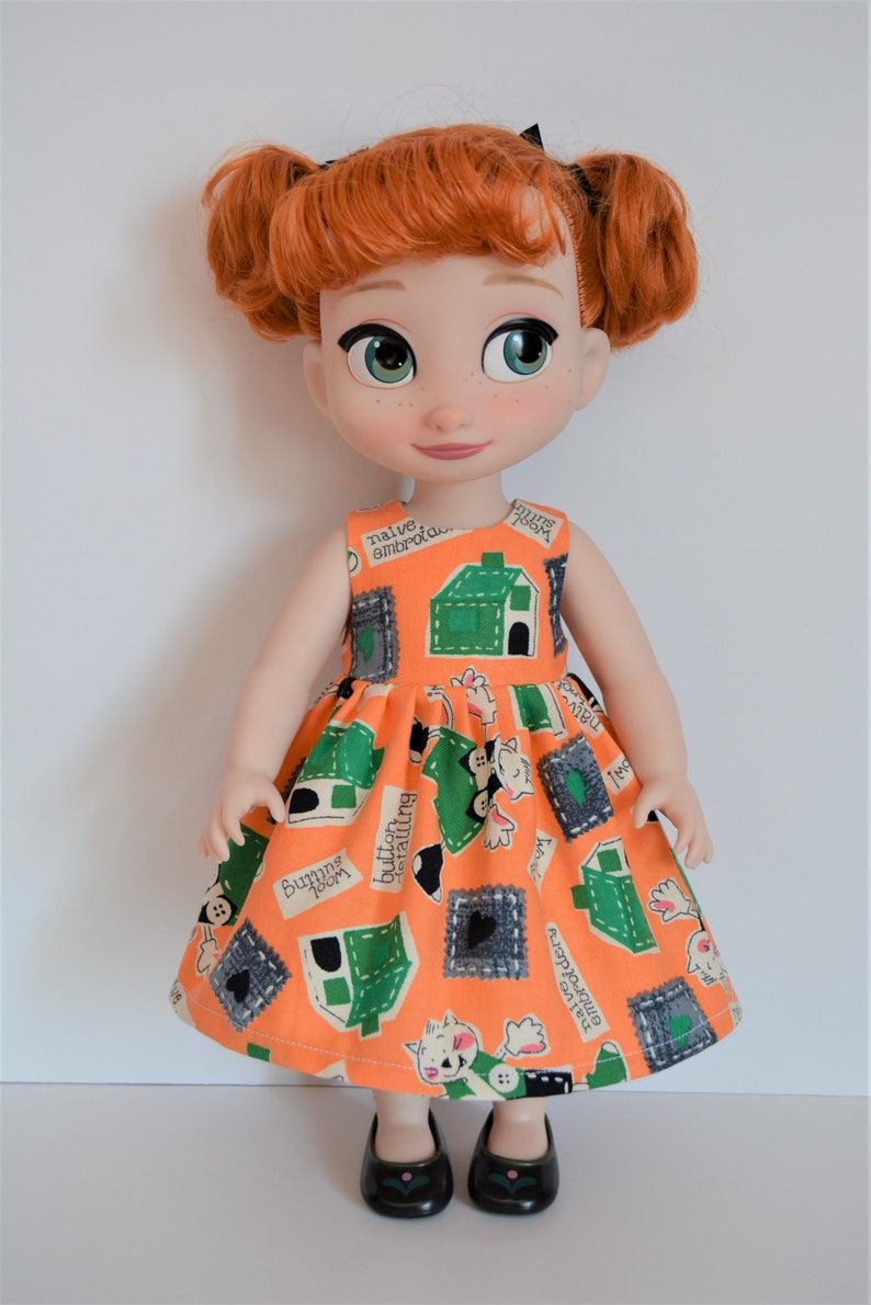 May include: A red-haired doll wearing a sleeveless orange dress with a green and white print. The dress has a variety of text on it, including "naive amorous", "buttons falling", and "funny little house". The doll is wearing black shoes.